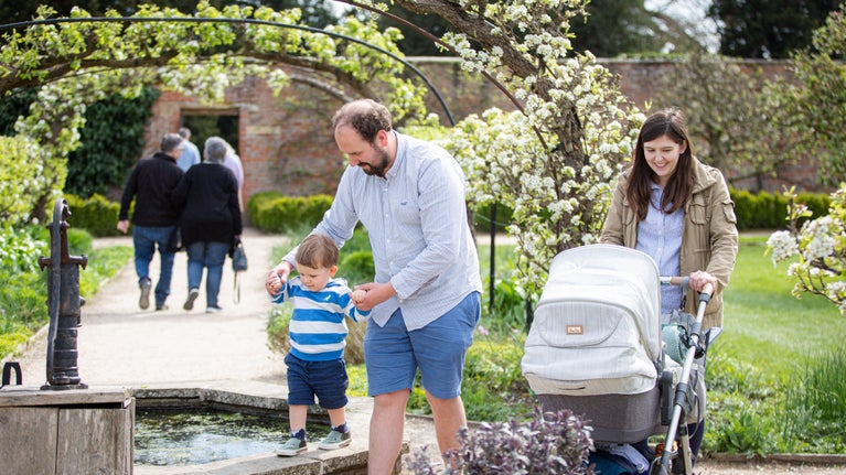 Man holding a young boys hands as he walks on a small pond wall, woman with a pram, walking under a blossoming arch